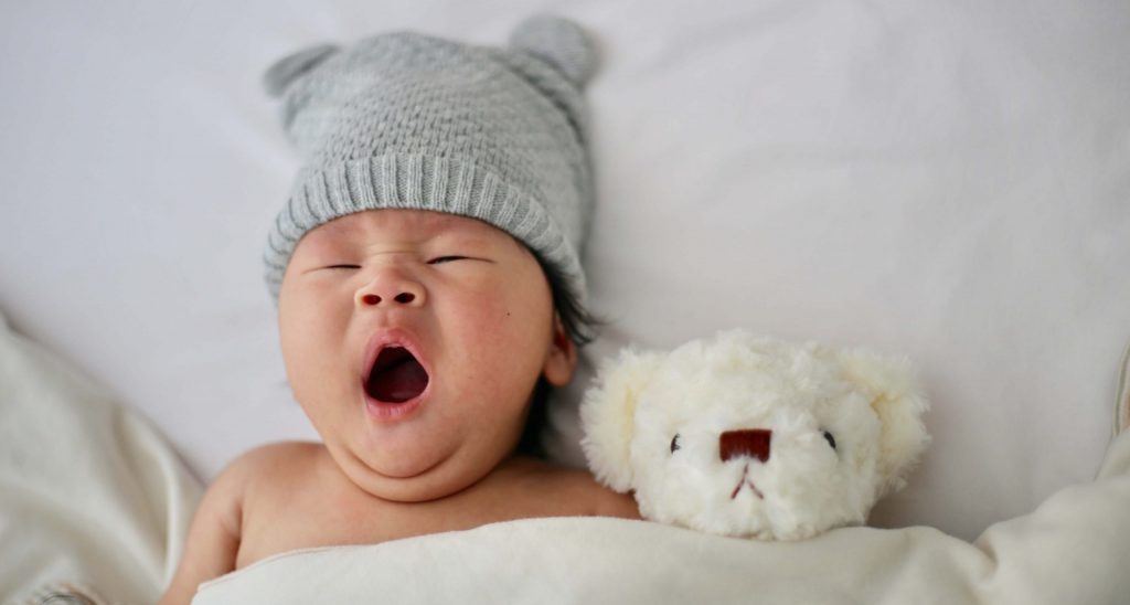 Ein Baby, das im Bett liegt und gähnt, neben sich einen weißen Kuschebären